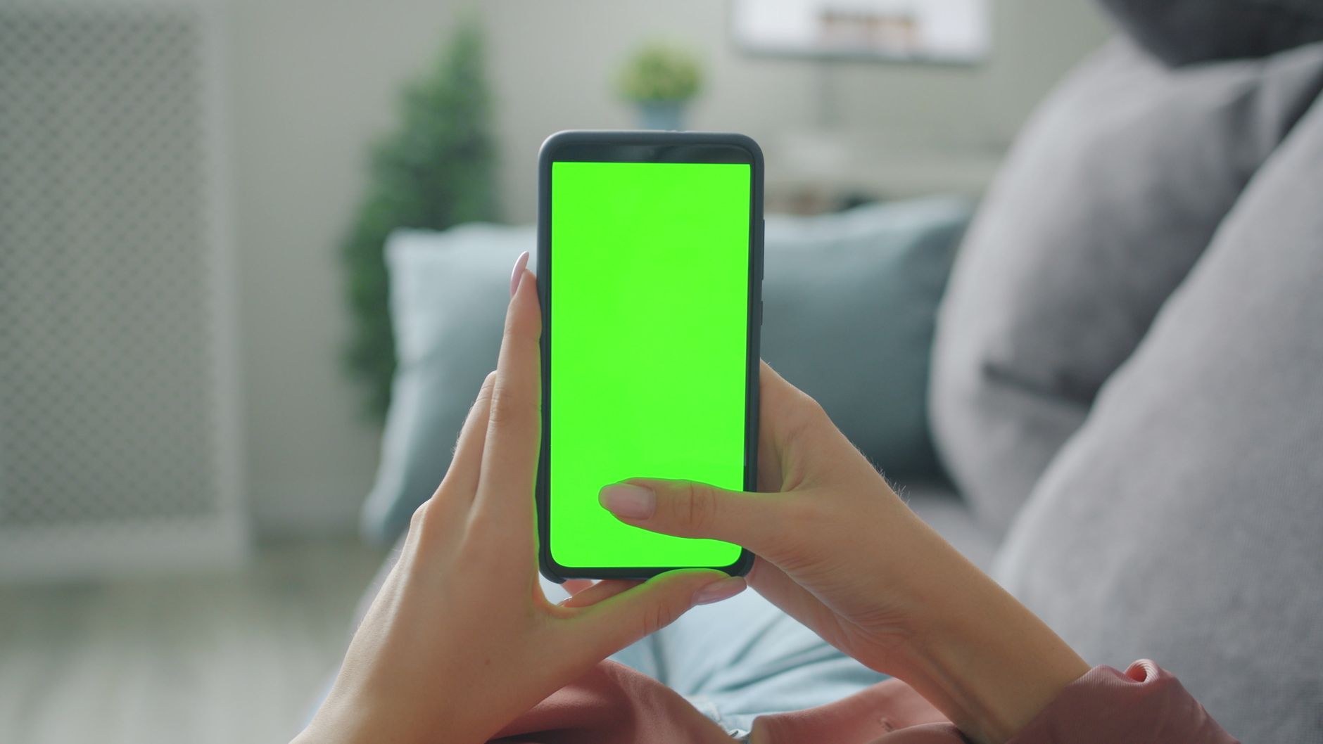 Duas mãos seguram um telemóvel com o ecrã verde, numa sala com sofá e almofadas.